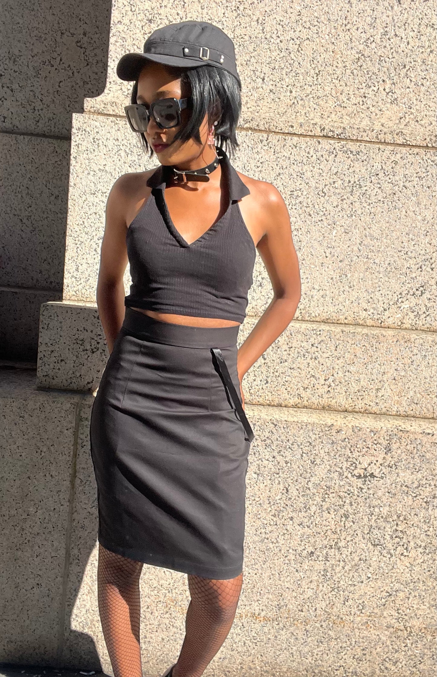 A model wearing KimYoung's knee-length black pencil skirt with KimYoung's signature ribbon chain in black, paired with a black top and black heels. The model is posing in front of a metal gate. The model is standing in front of a textured beige wall.