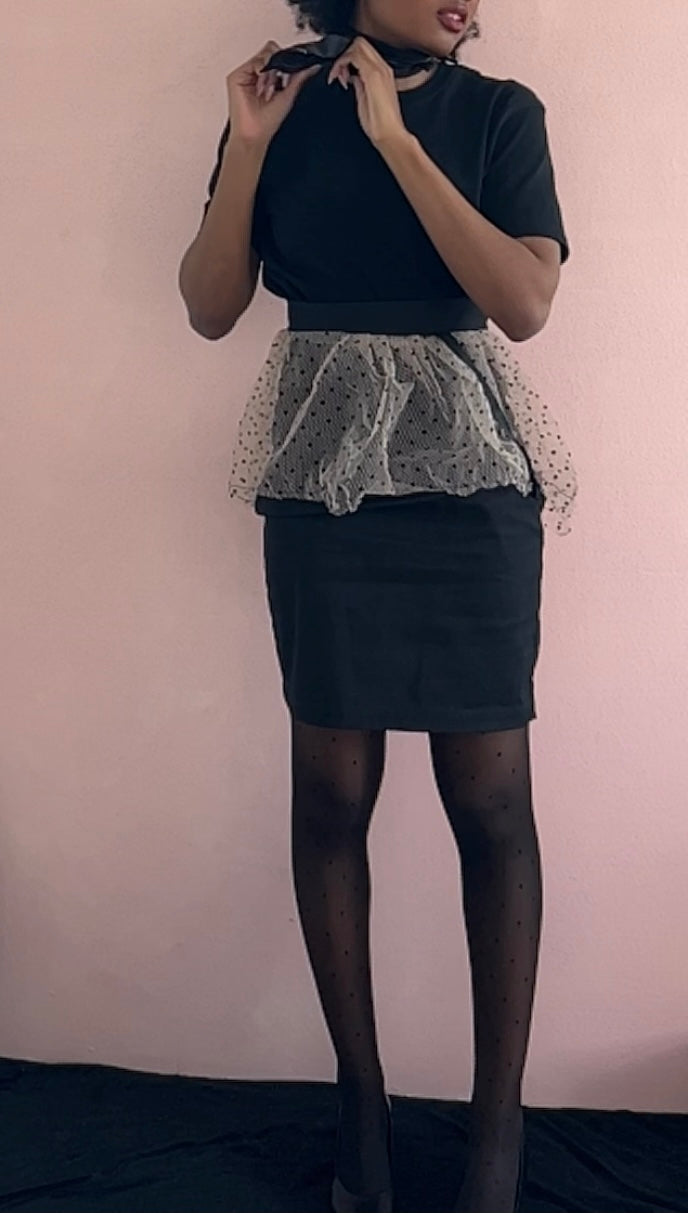 A model wearing KimYoung's knee-length black pencil skirt with a pink peplum peplum and black polka dots with KimYoung's signature black ribbon chain with a black bow with pearl on it, paired with a black top and black heels. The model is posing on the street in front of a support beam on the street.
