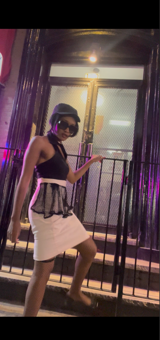 A model wearing KimYoung's knee-length pencil skirt with a black fishnet peplum and KimYoung's signature black ribbon chain with a black bow with pearl on it, paired with a black top and black heels. The model is standing in front of a metal gate.