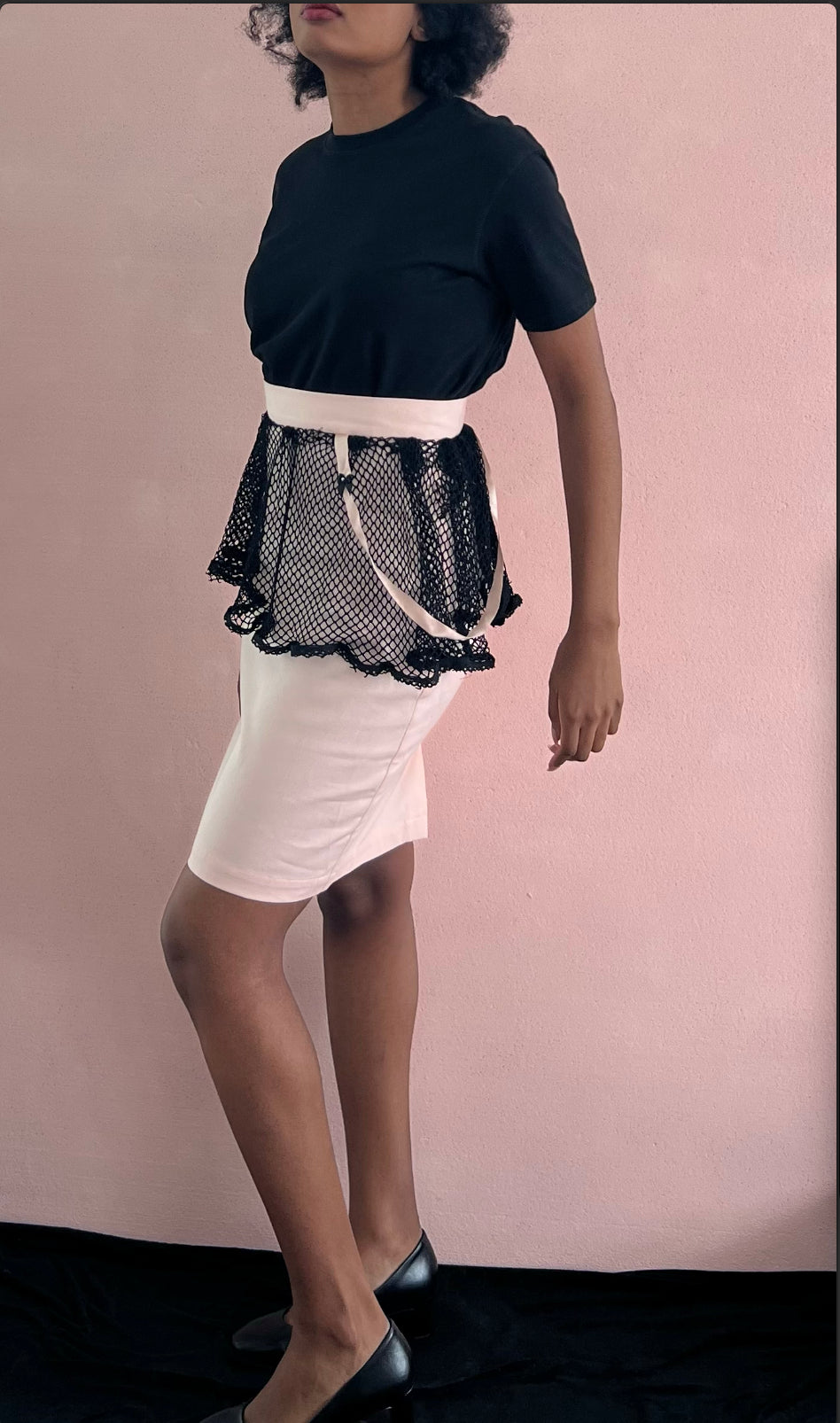 A model wearing KimYoung's knee-length pencil skirt with a black fishnet peplum and KimYoung's signature black ribbon chain with a black bow with pearl on it, paired with a black top and black heels.
