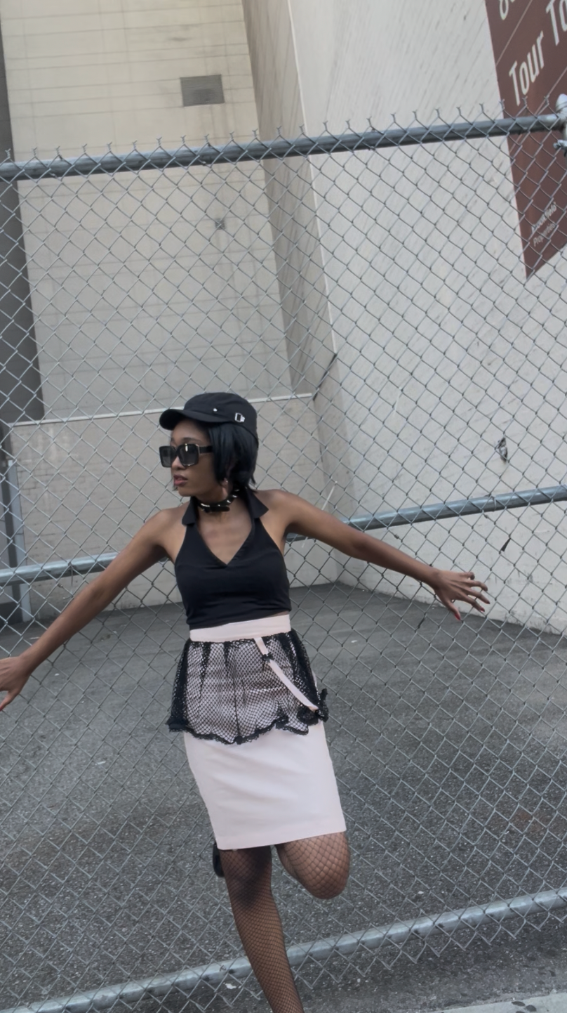 A model wearing KimYoung's knee-length pencil skirt with a black fishnet peplum and KimYoung's signature black ribbon chain with a black bow with pearl on it, paired with a black top and black heels. The models is posing in front of a metal chain link fence.