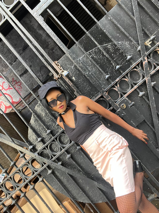 A model wearing KimYoung's knee-length pink pencil skirt with KimYoung's signature ribbon chain in pink, paired with a black top and black heels. The model is posing in front of a metal gate.