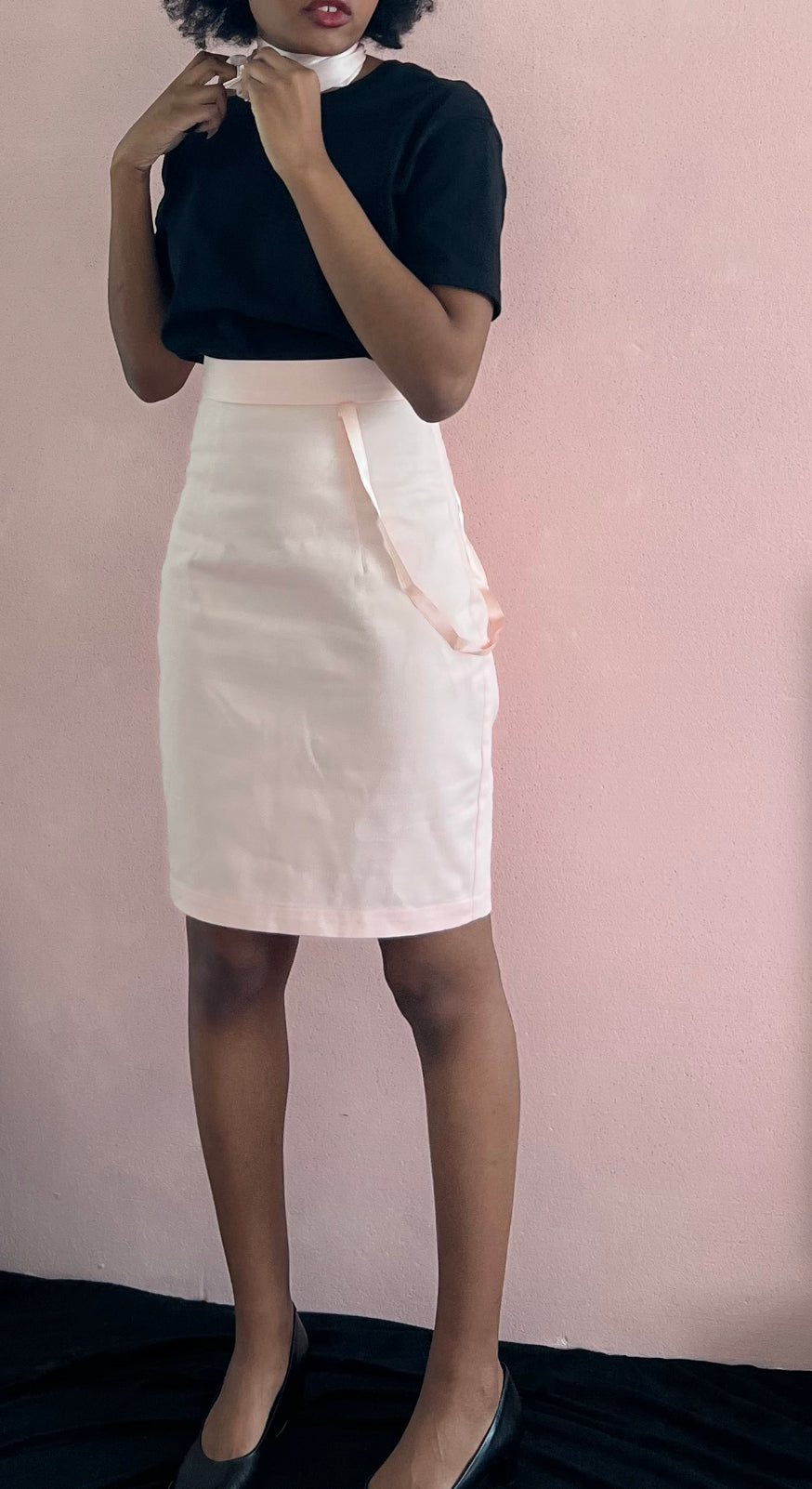 A model wearing KimYoung's knee-length pink pencil skirt with KimYoung's signature ribbon chain in pink, paired with a black top and black heels.