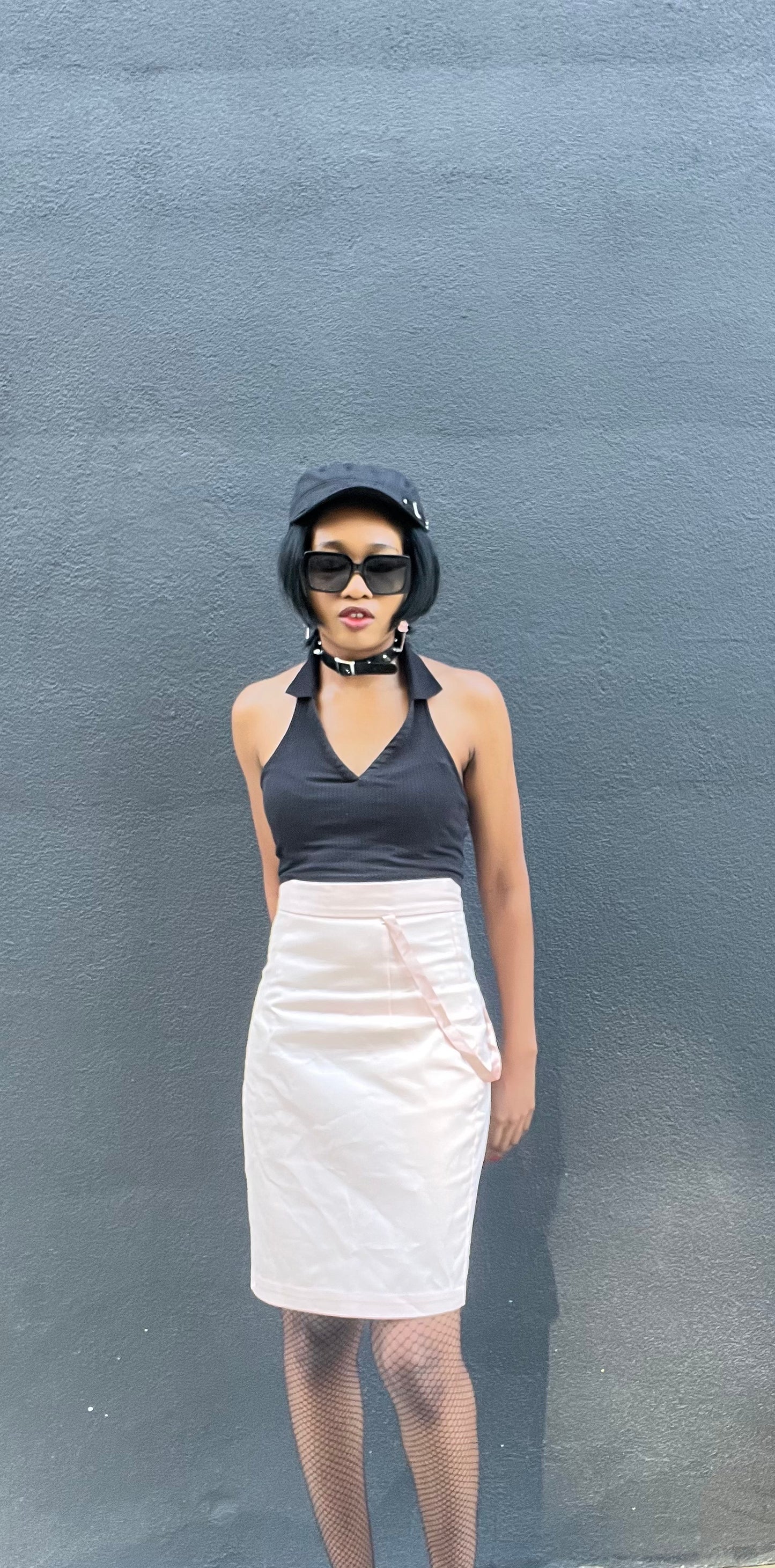 A model wearing KimYoung's knee-length pink pencil skirt with KimYoung's signature ribbon chain in pink, paired with a black top and black heels. The model is posing in front of a metal gate. The model is posing in front of a black wall.