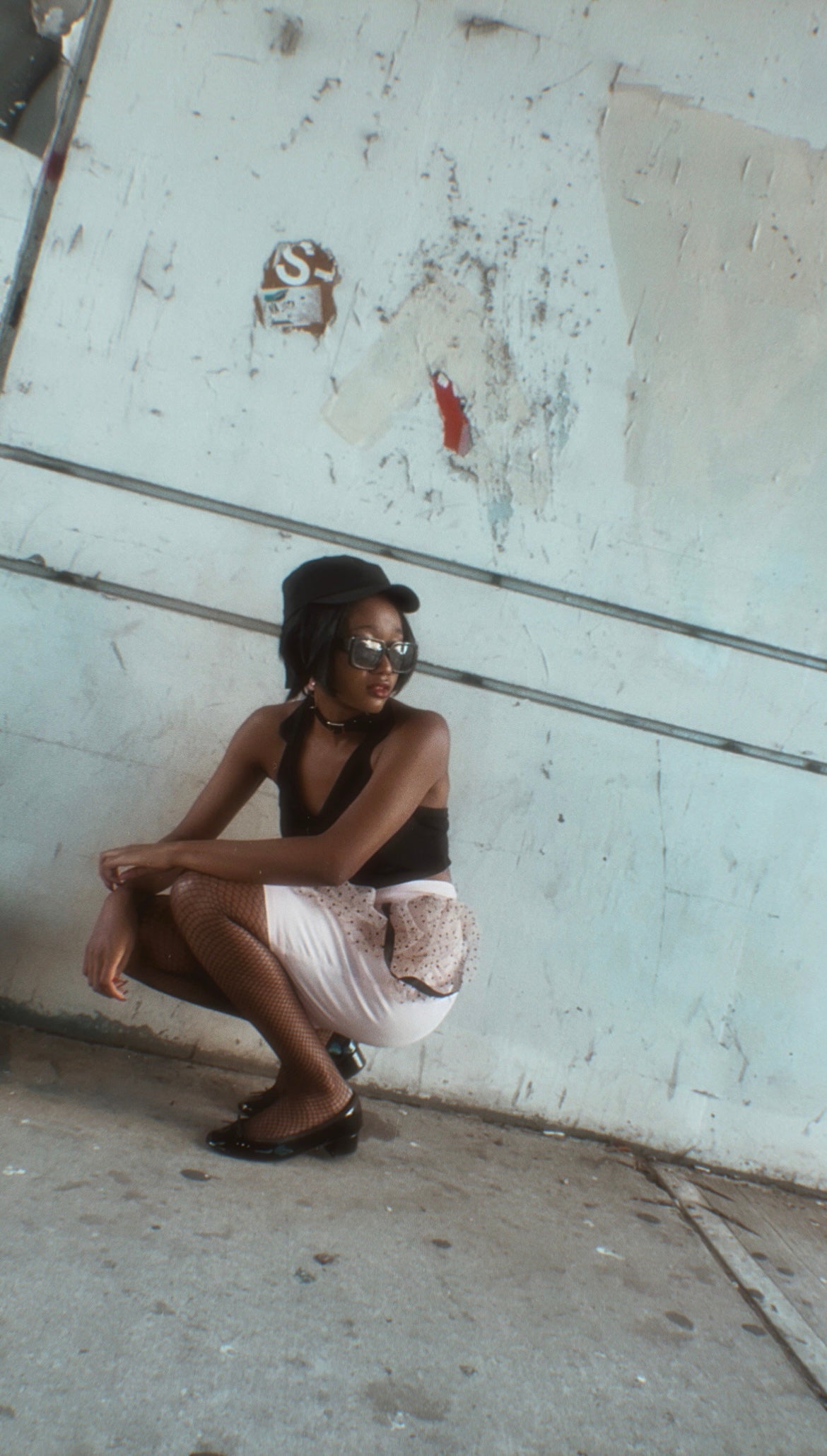 Woman squatting against a textured wall in an urban setting wearing KimYoung's pink polka pencil skirt with signature ribbon chain