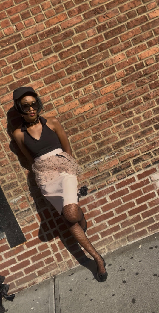 A model wearing KimYoung's knee-length pencil skirt with a pink polka dot peplum and KimYoung's signature black ribbon chain, paired with fishnet tights, a black top and black heels against a brick wall. 