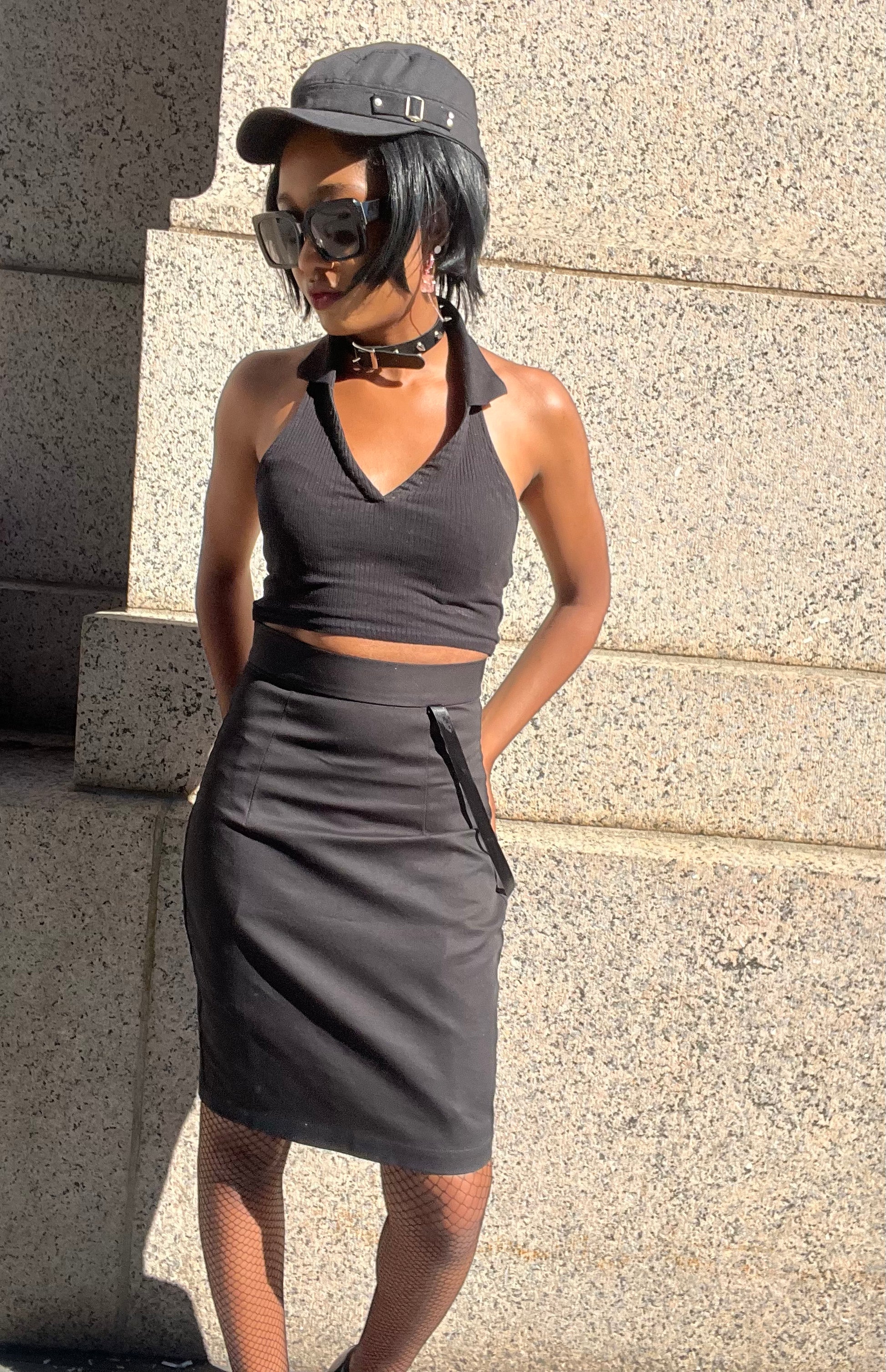 A model wearing KimYoung's knee-length black pencil skirt with KimYoung's signature  ribbon chain in black, paired with a black top and black heels. The model is posing in front of a metal gate. The model is standing in front of a textured beige wall. 