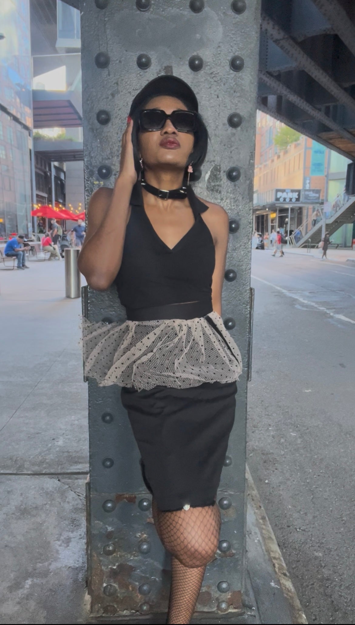 A model wearing KimYoung's knee-length black pencil skirt with a pink peplum peplum and black polka dots with KimYoung's signature black ribbon chain with a black bow with pearl on it, paired with a black top and black heels. The model is posing on the street in front of a support beam on the street. 