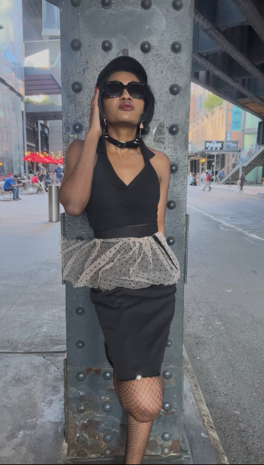 A model wearing KimYoung's knee-length black pencil skirt with a pink peplum peplum and black polka dots with KimYoung's signature black ribbon chain with a black bow with pearl on it, paired with a black top and black heels. The model is posing on the street in front of a support beam on the street. 