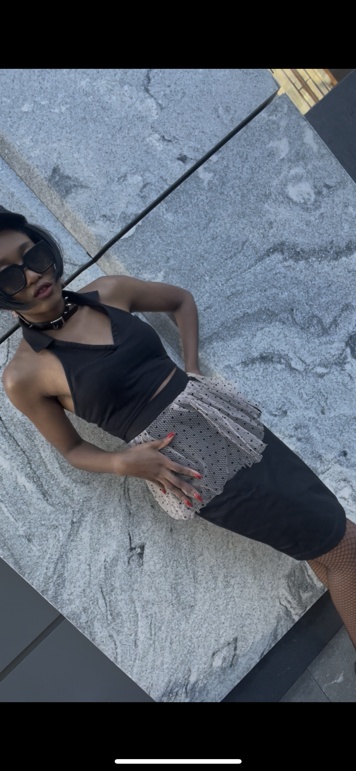 A model wearing KimYoung's knee-length black pencil skirt with a pink peplum peplum and black polka dots with KimYoung's signature black ribbon chain with a black bow with pearl on it, paired with a black top and black heels. The model is posing in front of a wall with a grey granite marbled design. 